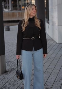 Ξανθιά γυναίκα με μακριά μαλλιά στέκεται σε ξύλινο διάδρομο φορώντας μαύρο σακάκι με ζώνη, ανοιχτό μπλε τζιν παντελόνι με φαρδιά μπατζάκια και κρατώντας μια μαύρη τσάντα χειρός.