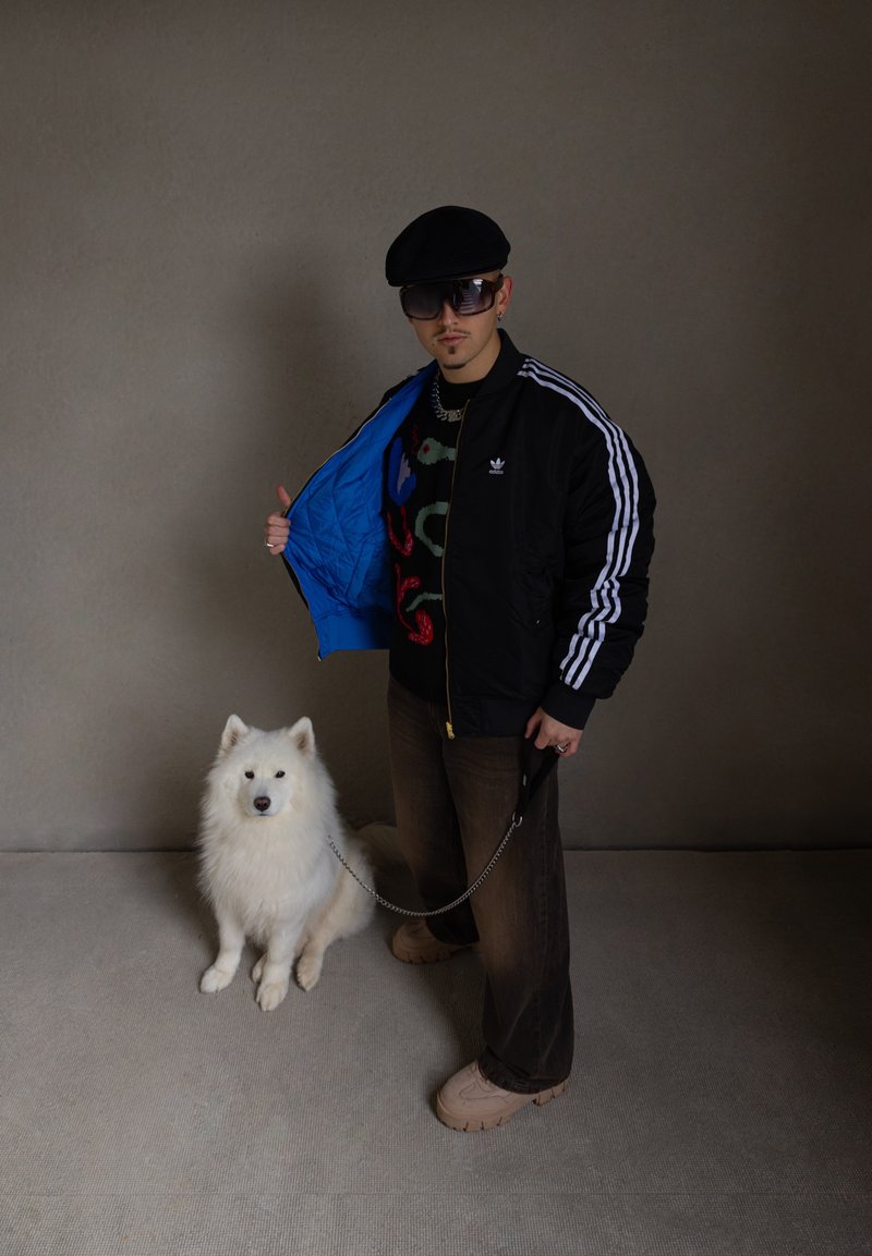 Μαύρο bomber τζάκετ Adidas με μπλε επένδυση, πολύχρωμο πουλόβερ με σχέδια, καφέ παντελόνι wide-leg, μπεζ παπούτσια και έναν λευκό Σαμόγιεντ δίπλα.