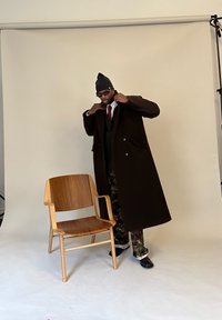 Muž upravuje tmavý dlouhý kabát vedle dřevěné židle, má na sobě černou čepici, sluneční brýle, červenou kravatu a maskáčové kalhoty ve studiu s béžovým pozadím.