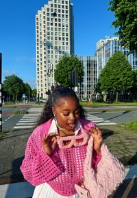 Rožā adīta krostopēde ar platām piedurknēm, balta blūze un atbilstoša pūkaina rožā soma. Tur rokās pūkaini rožā saulesbrilles.