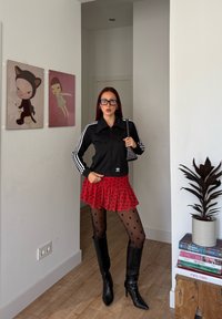 Kvinne med langt rødt hår iført svart Adidas-jakke, rød prikkete skjørt, svarte prikkete strømpebukser, støvler til knærne og briller, stående innendørs.