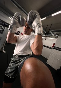 Sudraba boksa cimdi pacelti augšā, tiem tērpā cilvēkam baltā krekla un melnā šortos, ar tuvplānu uz ceļa priekšplānā.