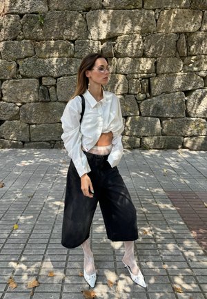 White cropped button-up shirt, black wide-leg trousers, white heels, sheer patterned tights. Stone wall backdrop with shadows.