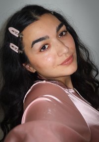 Selbstporträt einer Person mit langen, dunklen, welligen Haaren, die ein hellrosa Satinoberteil trägt. Haarklammern in einem ähnlichen Farbton unterstreichen die Frisur.