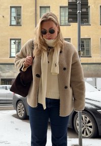 Casaco de fleece bege com botões pretos, cachecol creme, suéter creme e jeans azuis. Bolsa castanha. Rua nevada e carros ao fundo.