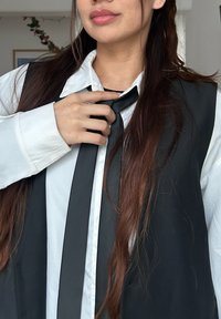 Cravate noire portée sur une chemise blanche avec un col. La cravate est texturée, et la chemise a des manches longues retroussées. Les cheveux sont longs et foncés.