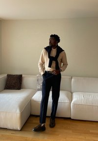 Bărbat stând într-o cameră lângă o canapea bej, purtând o cămașă bej deschis, pantaloni închiși la culoare, pantofi maro și un pulover așezat pe umeri.