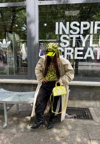 Hoodie z vzorcem leoparda z zelenimi detajli, bež trench coat, črne široke hlače, črne konične škornje in majhna rumena torbica.