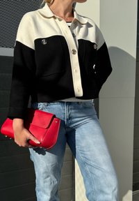 Μαύρο και άσπρο μπουφάν με σχέδιο κουμπώματος, δύο μπροστινές τσέπες και ασημί λεπτομέρειες. Κρατώντας μια κόκκινη δερμάτινη τσάντα, σε συνδυασμό με ανοιχτό μπλε τζιν.