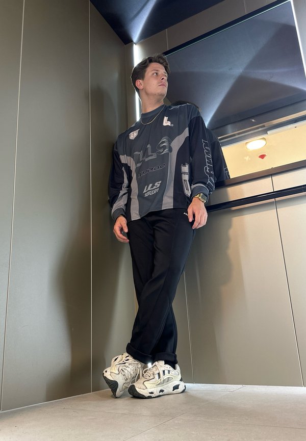 Camisa gráfica de manga larga en negro y gris, pantalones negros y zapatillas blancas con acentos negros. De pie en un interior moderno con paredes elegantes.