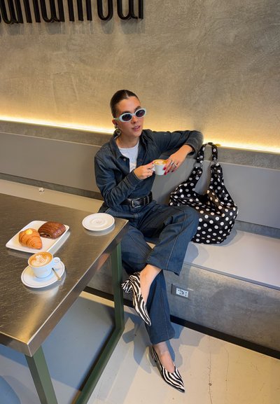 Mujer con atuendo de mezclilla y zapatos con patrón de cebra sentada en una cafetería sosteniendo una taza de café, con croissants y un latte en la mesa cercana.