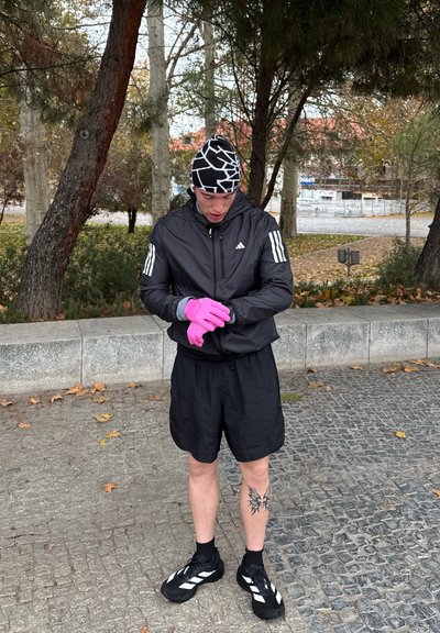 Hombre con ropa deportiva negra, guantes rosas y gorra estampada mira su reloj mientras está de pie en un sendero con árboles y hojas de otoño cerca.