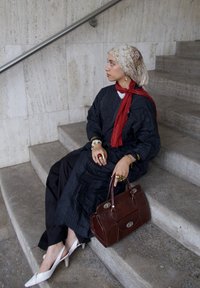 Roba lunga a strisce nere, sciarpa rossa, foulard a motivo, gioielli d'oro, borsa marrone e décolleté bianchi a punta. Seduta su scale in pietra.