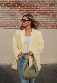 Υπερμεγέθης κίτρινος σακάκι, λευκή αμάνικη μπλούζα και ανοιχτό μπλε τζιν. Κρατάει μια υφασμάτινη τσάντα σε ελιά. Το φόντο περιλαμβάνει έναν τοίχο από τούβλα.