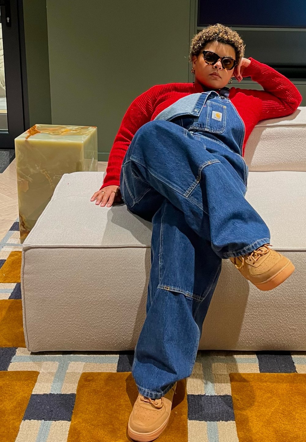 Τζιν σαλοπέτα σε μπλε με φαρδιά γραμμή, σε συνδυασμό με κόκκινη ριγωτή μπλούζα και καφέ αθλητικά παπούτσια. Καθισμένη σε έναν ανοιχτόχρωμο καναπέ.