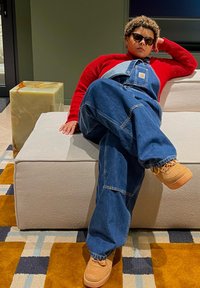 Τζιν σαλοπέτα σε μπλε με φαρδιά γραμμή, σε συνδυασμό με κόκκινη ριγωτή μπλούζα και καφέ αθλητικά παπούτσια. Καθισμένη σε έναν ανοιχτόχρωμο καναπέ.