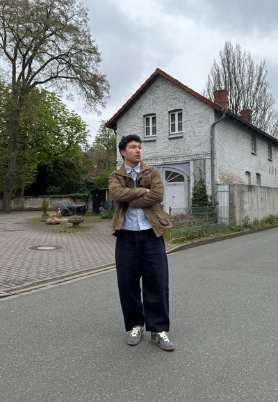 Joven con chaqueta marrón y pantalones oscuros, de pie con los brazos cruzados en una calle gris cerca de una casa blanca de ladrillo y árboles bajo un cielo nublado.