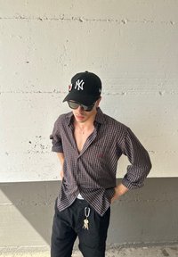 Camicia a quadri con colletto aperto, con un motivo in colori scuri, indossata sopra pantaloni neri. Berretto nero con logo; occhiali da sole completano il look.