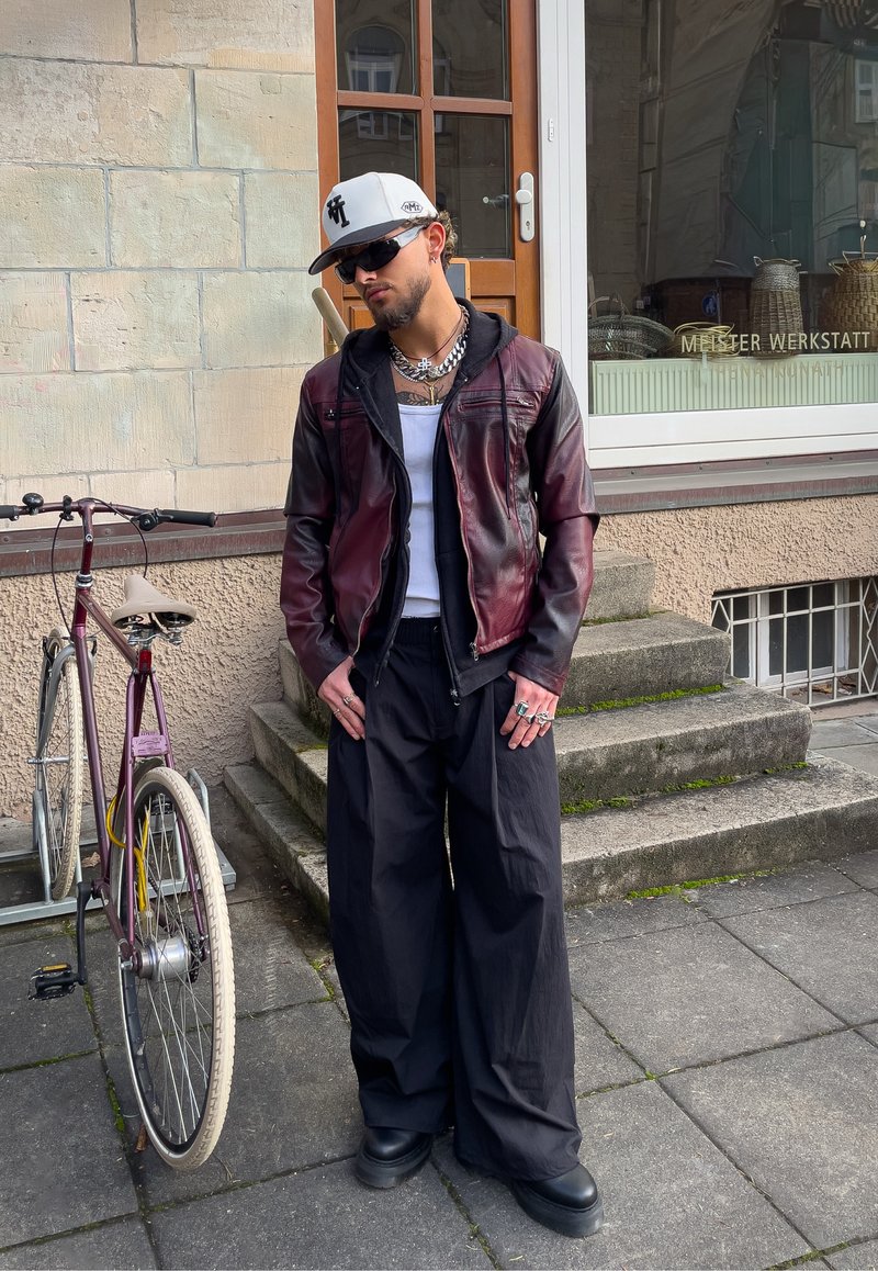 Personne portant une veste en cuir bordeaux sur une chemise blanche, un pantalon noir large et des bottes noires, se tenant à côté d'un vélo violet.