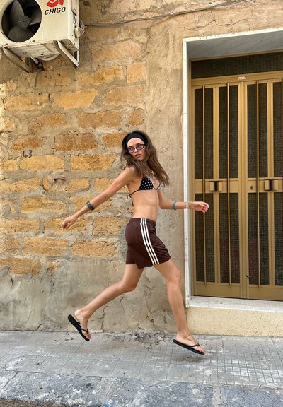 Una persona con un top de bikini de lunares y pantalones cortos deportivos marrones con rayas blancas camina por una acera junto a una pared de piedra texturizada y una puerta.