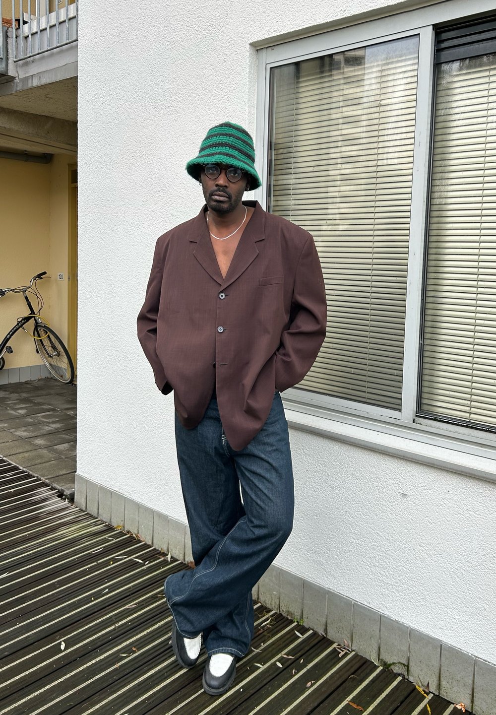 Blazer castanho oversized com três botões, combinado com jeans justos de flare azul-escuro e sapatos preto e branco. Chapéu de balde listrado verde e óculos.