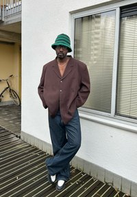 Blazer castanho oversized com três botões, combinado com jeans justos de flare azul-escuro e sapatos preto e branco. Chapéu de balde listrado verde e óculos.