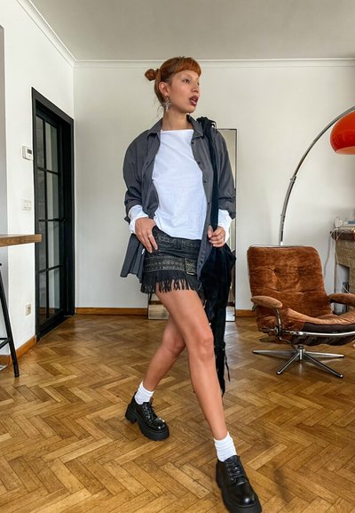 Mujer caminando en interior vistiendo falda negra con flecos, camisa blanca, chaqueta abierta a rayas, calcetines blancos, zapatos de plataforma negros y pendientes plateados.