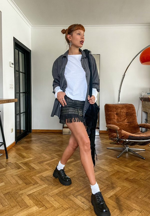 Frau, die in Innenräumen geht, trägt einen schwarzen Fransenrock, ein weißes Hemd, eine gestreifte offene Jacke, weiße Socken, schwarze Plateauschuhe und silberne Ohrringe.