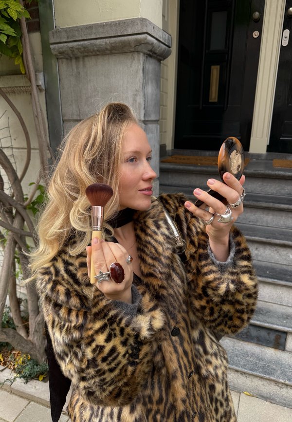 Kvinna med blont hår som bär en leopardmönstrad kappa applicerar smink med en borste medan hon håller i en kompakt spegel utanför en byggnad med stentrappor.