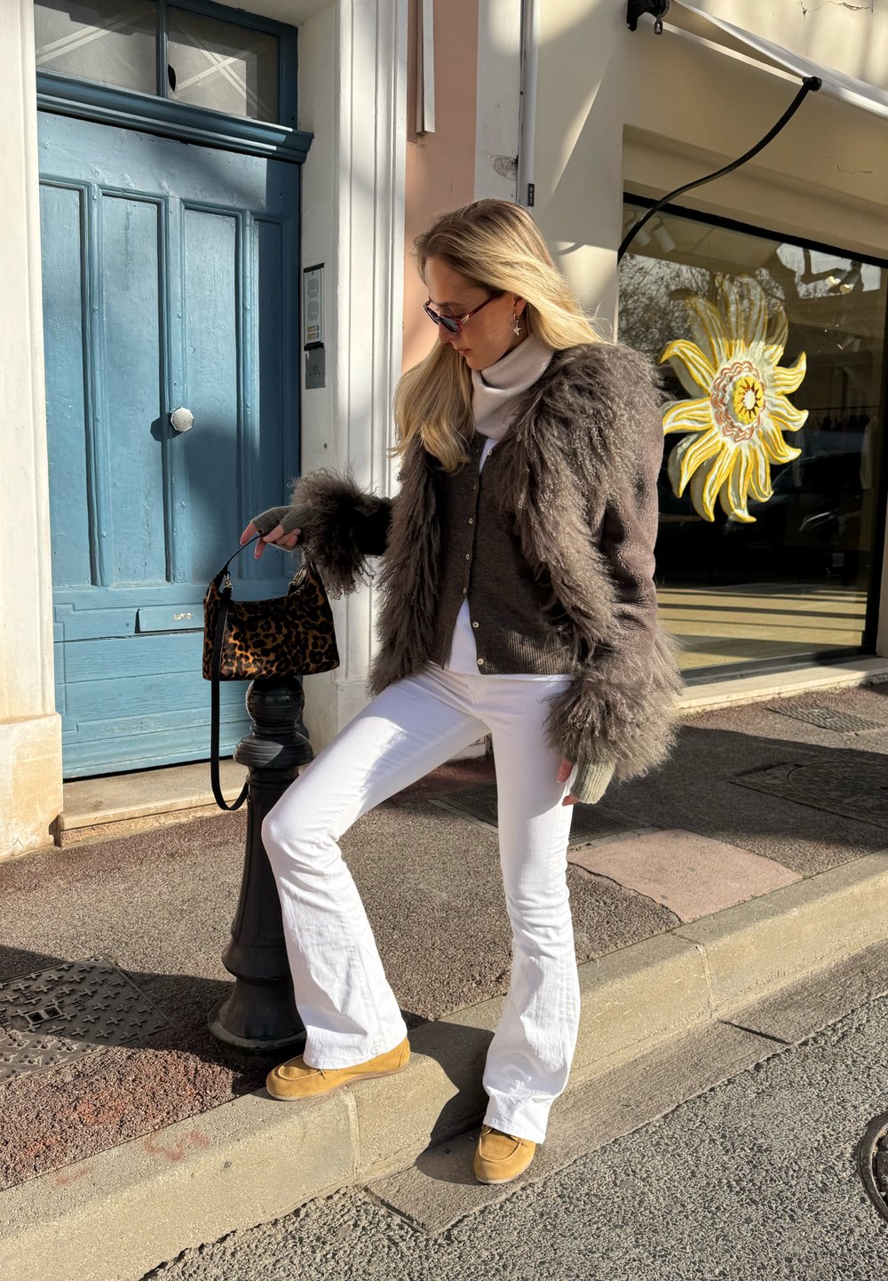 Femme en pantalon blanc et veste marron bordée de fourrure ajuste un sac à main à motif léopard sur un bollard de rue noir près d'une porte bleue.