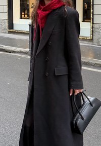 Čierny dvojradový vlnený kabát s dlhým siluetom, pod ním červená textúrovaná kapucňa a malá čierna kabelka s oblými rúčkami.