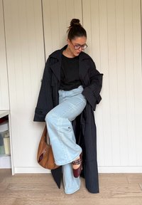 Dlhý čierny kabát s pásikmi, čierna košeľa, svetlomodré rozšírené džínsy, hnedá taška a bordové topánky. V pozadí drevená podlaha a biele obklady.