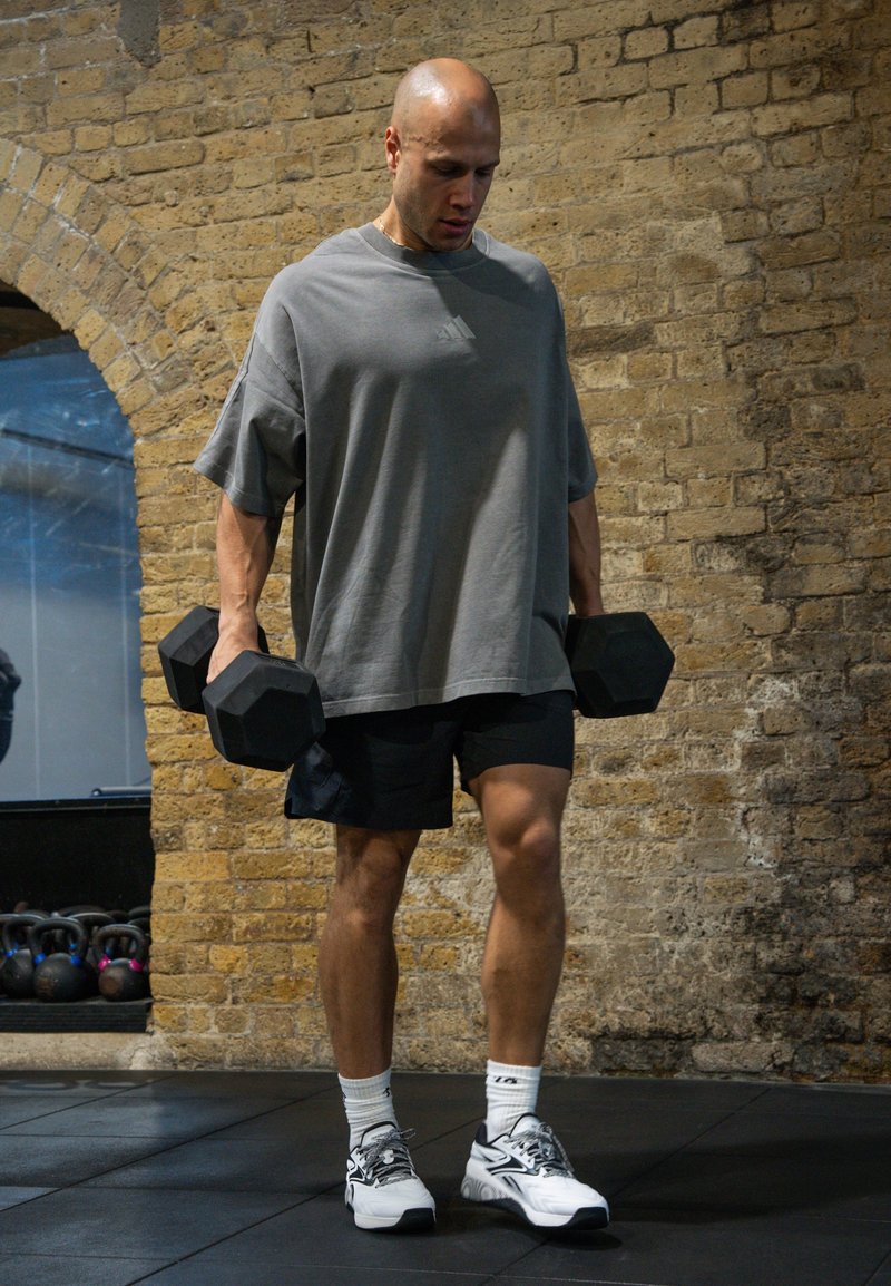 Homme athlétique en t-shirt gris et short noir tenant des haltères, debout sur le sol de la salle de sport contre un mur en briques, se préparant à s'exercer.