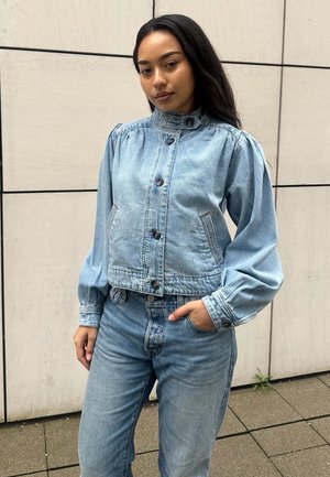 Mujer joven con cabello largo y oscuro que lleva una chaqueta vaquera azul claro con botones y vaqueros a juego, de pie contra una pared de azulejos beige.