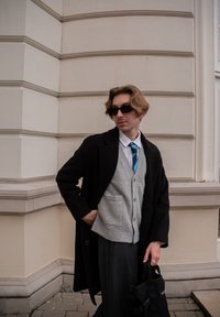 Personne portant un manteau noir par-dessus un pull gris, une chemise blanche et une cravate turquoise. Tenant un sac noir, se tenant contre un mur texturé.