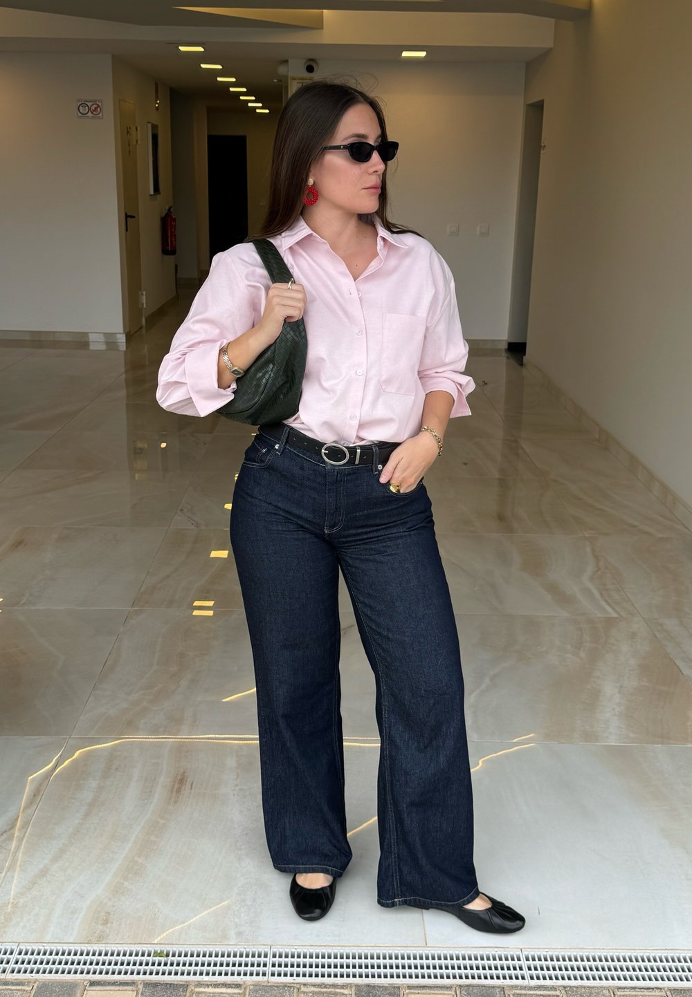 Svetlo roza košulja s dugim rukavima, temno modri zvonasti traperice, črne ravne cipele, drži teksturiranu zeleno torbico, nosi črne sončne očala in rdeče uhane.