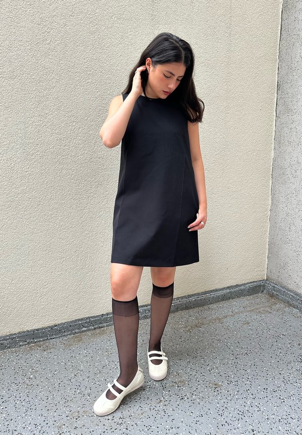 Mujer con un vestido negro sin mangas, medias negras transparentes hasta la rodilla y zapatos blancos, de pie sobre un suelo moteado cerca de una pared texturizada.