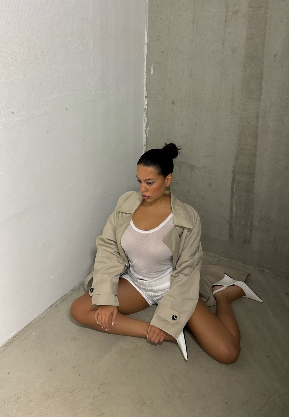 Casaco trench bege sobre uma blusa branca e calções brilhantes; modelo está sentada com cabelo escuro liso e usa saltos stiletto brancos.