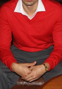 Roter Pullover mit V-Ausschnitt über einem weißen Hemd, kombiniert mit grauen Hosen. Hände liegen auf einer Holzoberfläche, ein goldener Armbanduhren ist sichtbar.