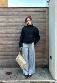 Sort dobbeltbrystet cropped jakke med bred krave, grå vidde bukser og beige håndtaske. Blank mørk sko med lyse accenter.