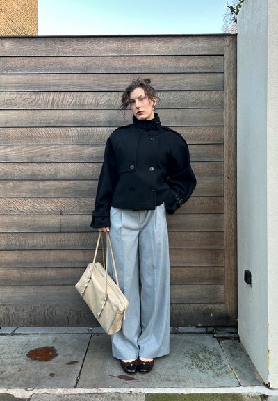 Chaqueta negra cruzada corta con cuello amplio, pantalones anchos grises y bolso beige. Zapatos oscuros brillantes con detalles claros.