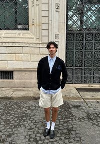 Gilet noir avec des boutons en bois sur une chemise bleu clair, associé à un short beige et des mocassins noirs, se tenant sur un pavé en pierre.