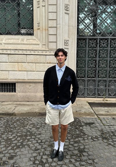 Cardigan negro con botones de madera sobre una camisa azul clara, combinado con pantalones cortos beige y mocasines negros, de pie sobre un pavimento de adoquines.