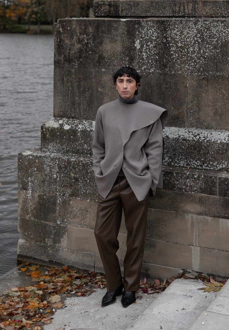 Personne aux cheveux bouclés portant une veste grise asymétrique et un pantalon marron, debout contre un mur en pierre près de l'eau et de feuilles d'automne tombées.