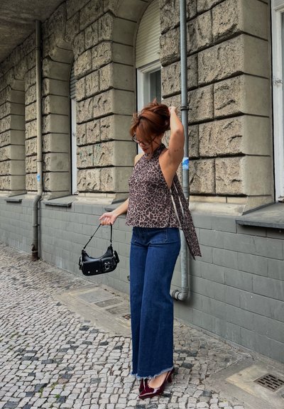Mujer con cabello rojo, top sin mangas con estampado de leopardo, vaqueros de pierna ancha y tacones burdeos sosteniendo un bolso negro en una calle adoquinada.