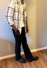 Pantalon noir, t-shirt blanc et chemise à manches longues à carreaux. La chemise présente des carreaux bleus et gris. Chaussures noires avec semelles texturées.