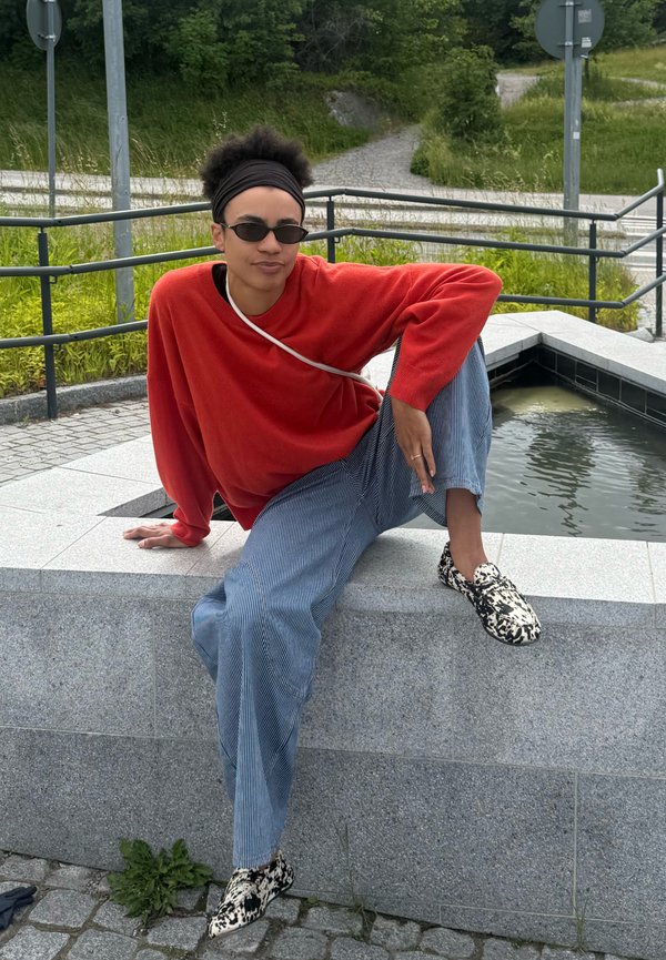 Personne portant un pull rouge, un pantalon à rayures et des chaussures à motifs assise sur un rebord en pierre près d'un petit élément d'eau extérieur.