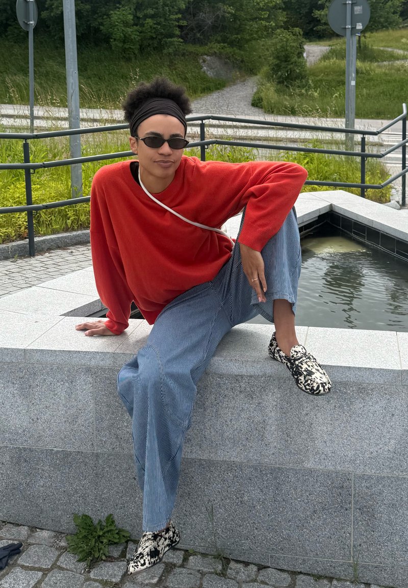 Personne portant un pull rouge, un pantalon rayé et des chaussures à motifs assise sur un rebord en pierre à côté d'un petit bassin d'eau à l'extérieur.