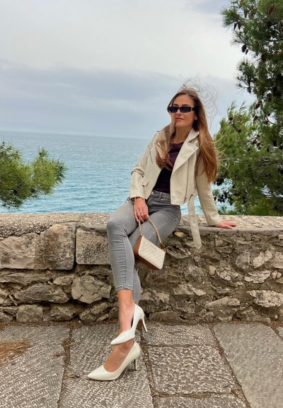 Una chaqueta corta de cuero crema sobre una blusa oscura, jeans ajustados grises, zapatos de tacón blanco con punta y un pequeño bolso descansando sobre un muro de piedra.