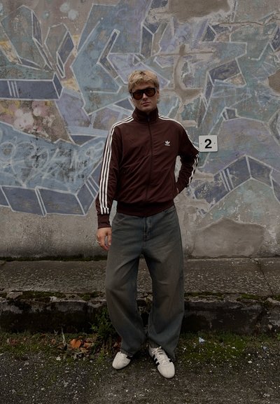 Chaqueta de chándal marrón con cremallera y rayas blancas, jeans gris sueltos y zapatillas blancas. El fondo presenta un mural abstracto colorido.